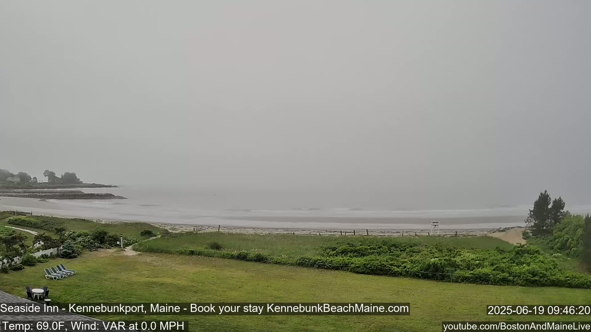 A foggy, overcast view shows a beach with a calm ocean, green grassy areas, and some trees in the distance.