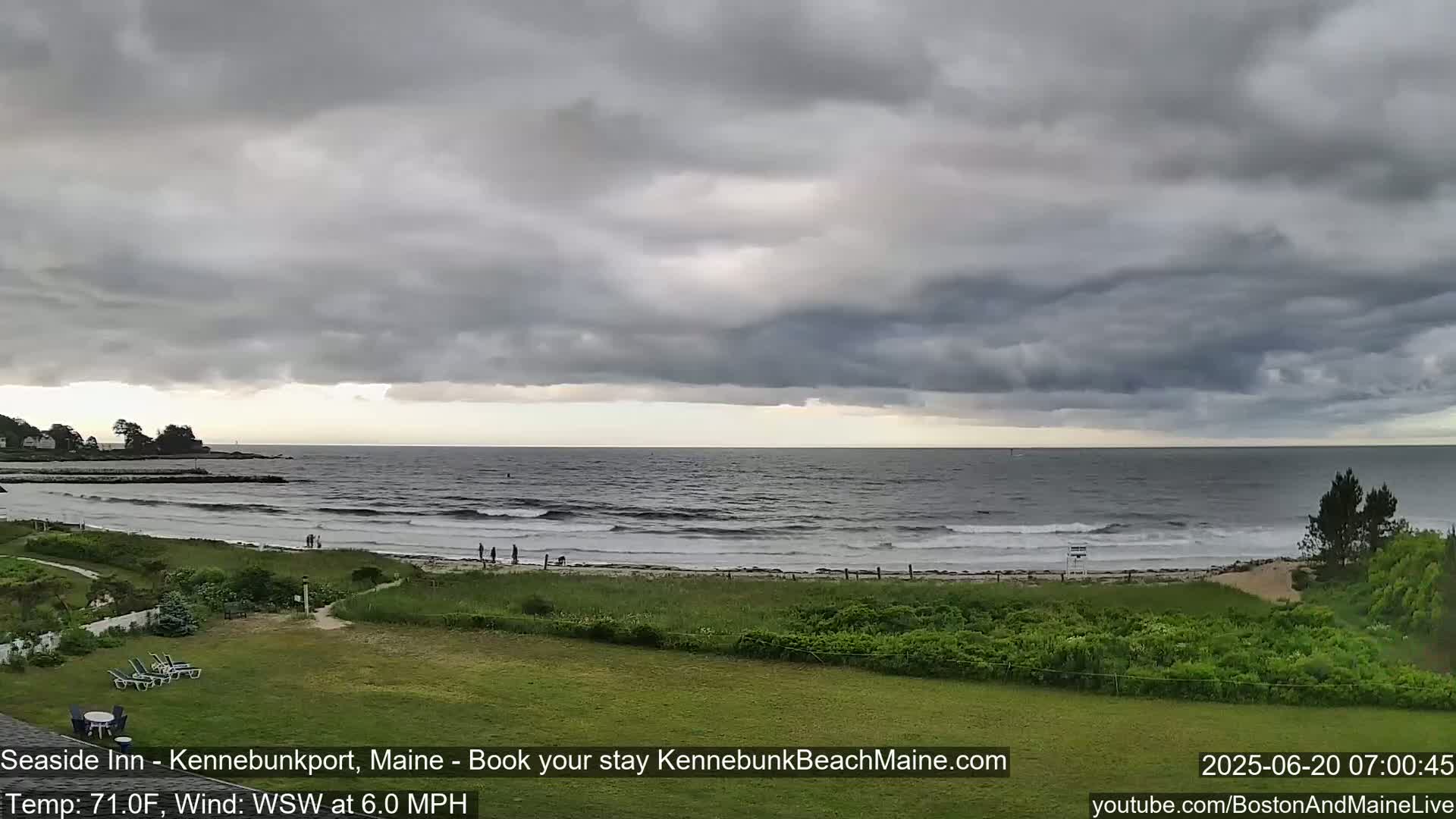 A beach scene under a gray, cloudy sky shows a calm ocean with small waves lapping the shore, where a few people are walking, and grassy areas with some vegetation.