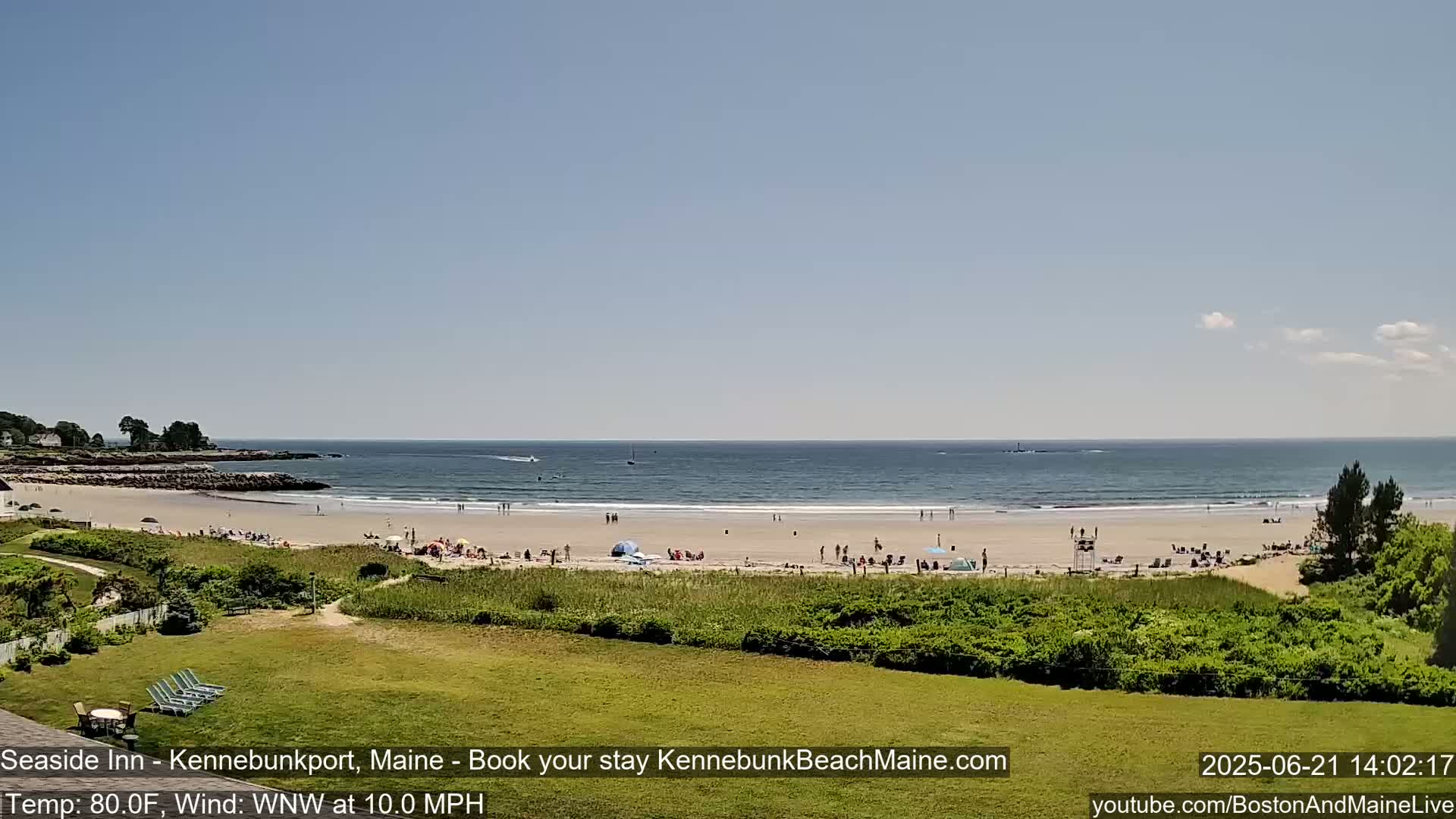 A sandy beach with scattered people on a sunny day, viewed from a grassy area overlooking the ocean with small waves.
