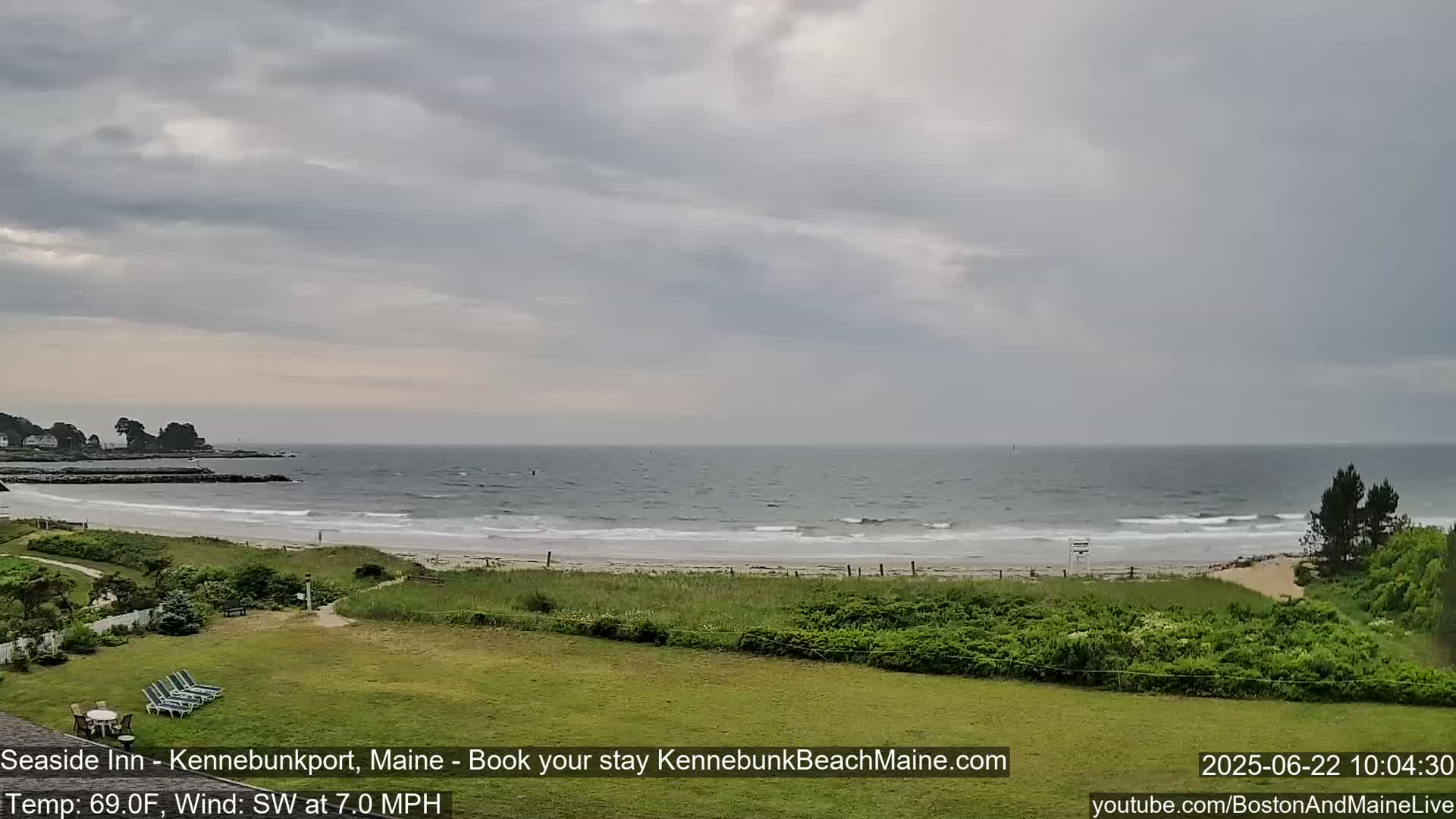 A view of a beach with gentle waves under an overcast sky, seen from a grassy area with lawn furniture.