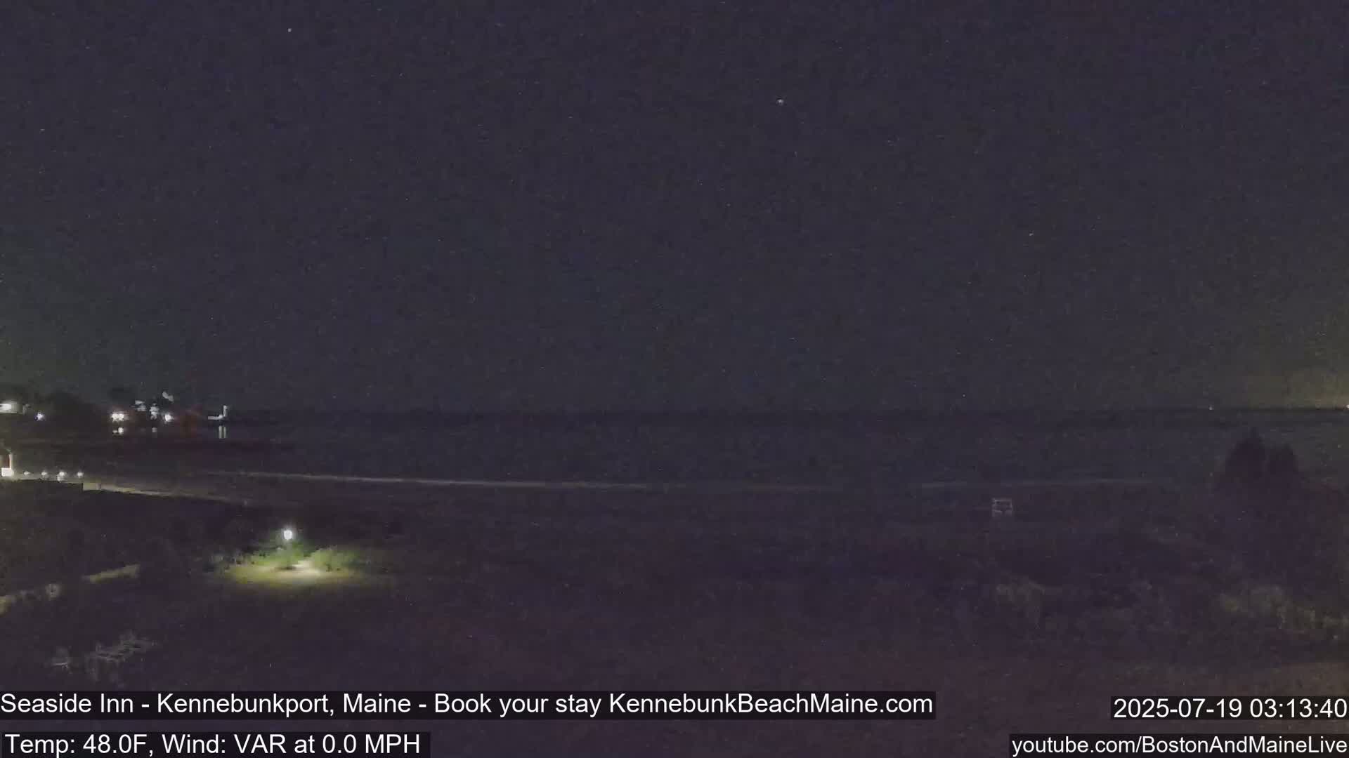 A nighttime coastal scene shows a dark ocean under a mostly clear sky, with faint lights visible in the distance and a small lighted area in the foreground.