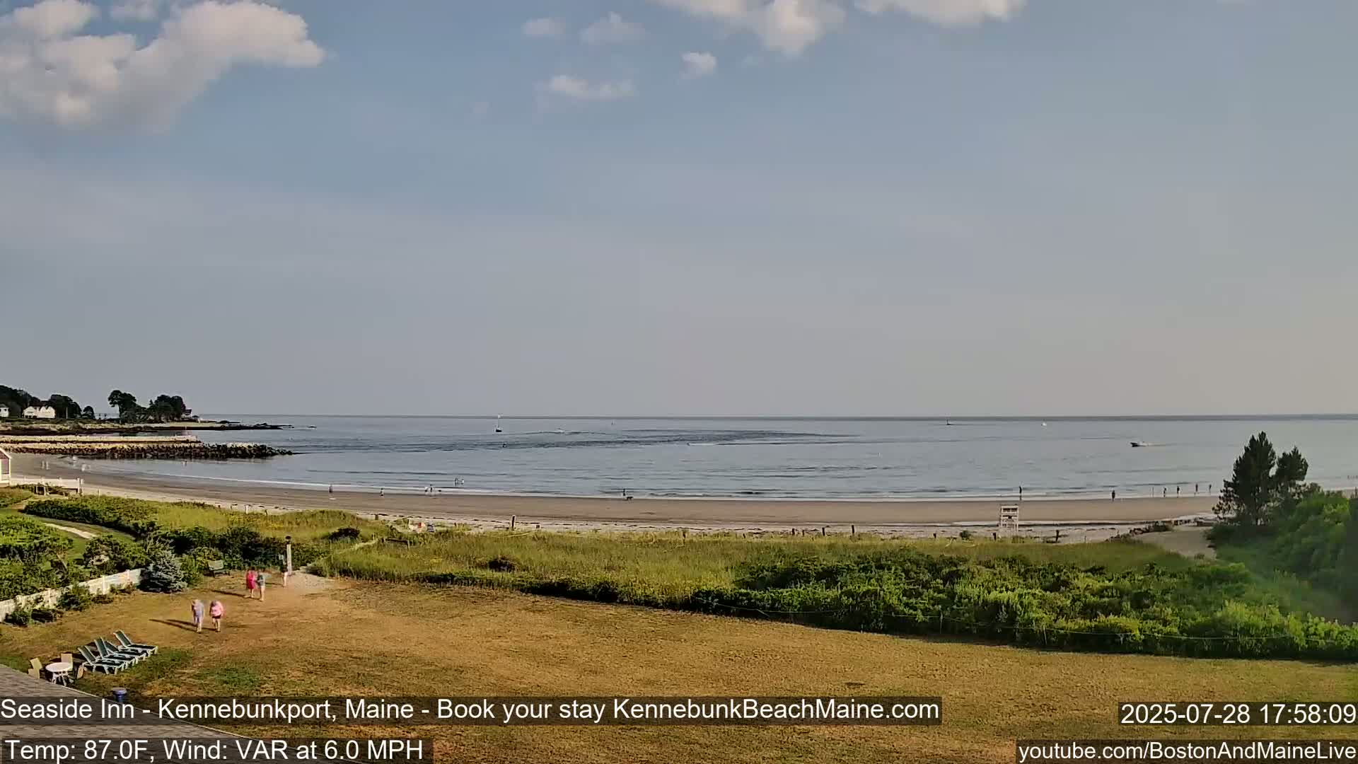 A sandy beach with a calm ocean under a partly cloudy sky is viewed from a grassy area with a few people walking.