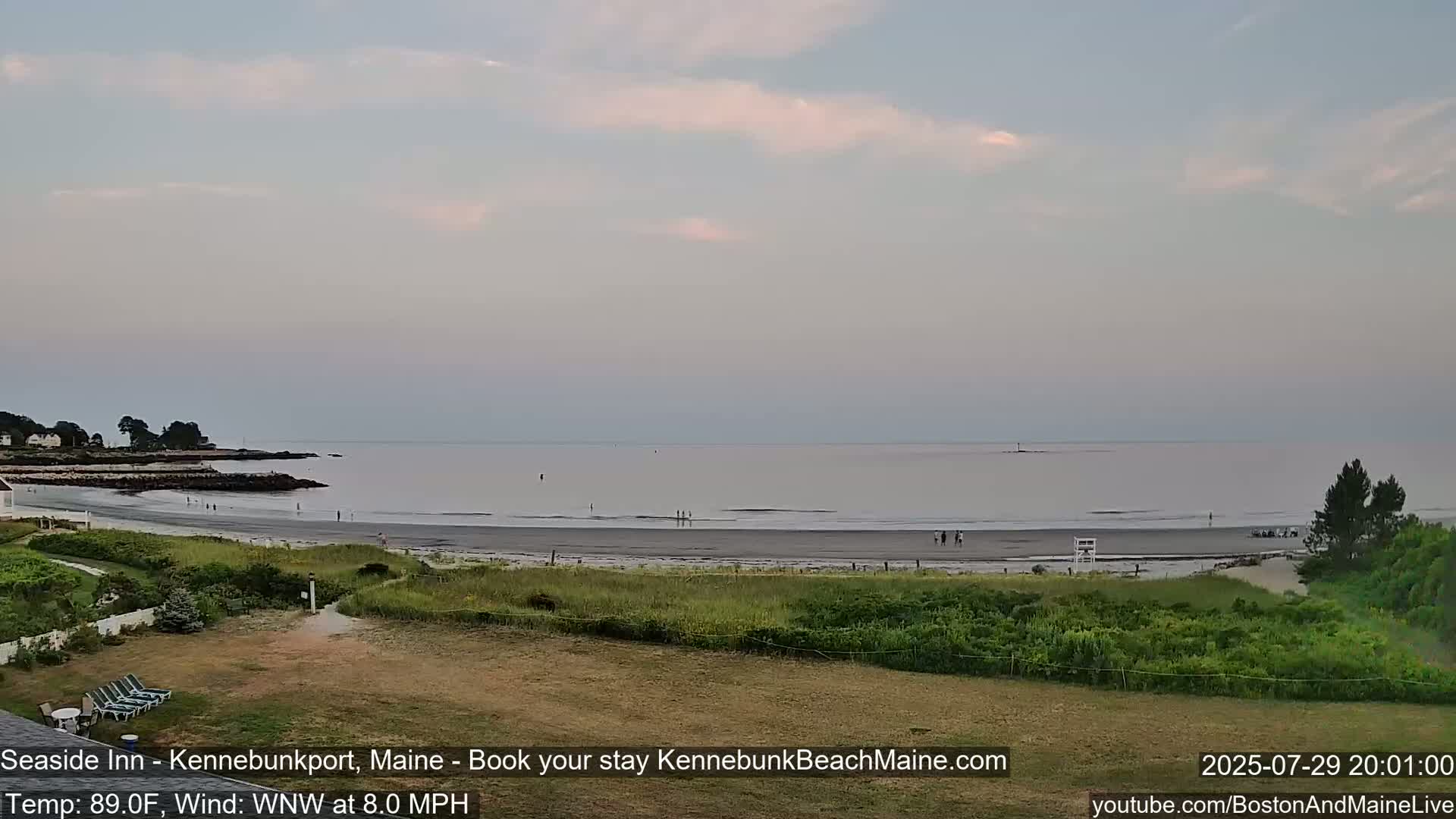 A sandy beach with a calm ocean under a partly cloudy sky is viewed from a grassy area with a few people walking.