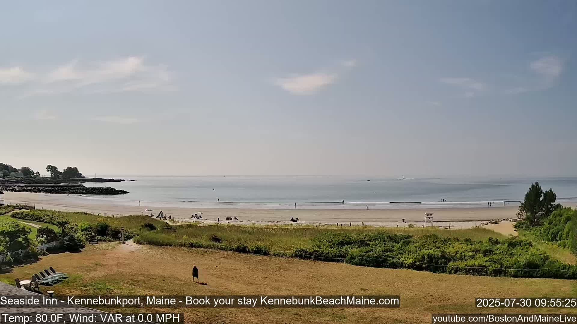 A sandy beach with a calm ocean under a partly cloudy sky is viewed from a grassy area with a few people walking.