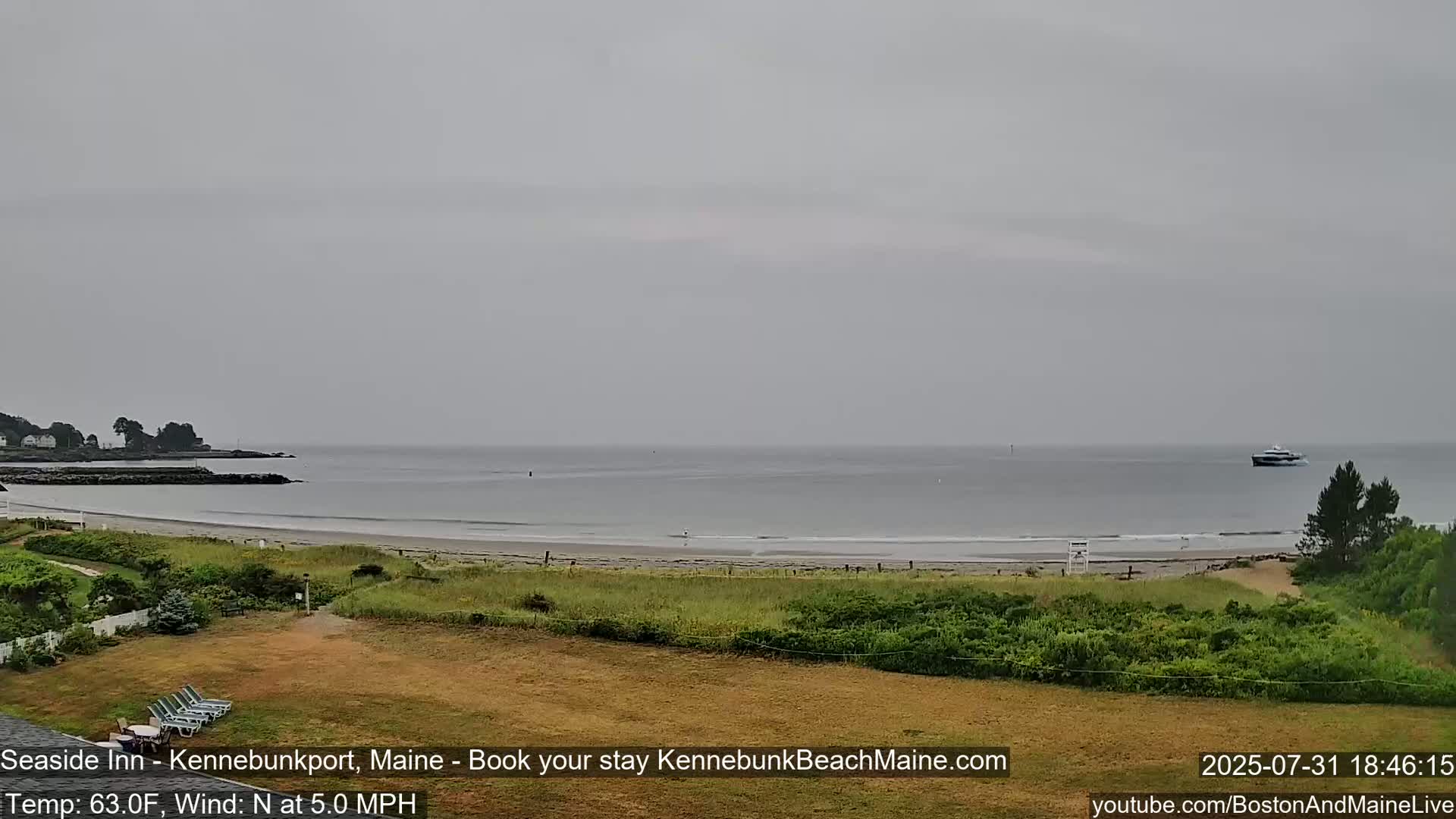 A beach with calm, gray water under an overcast sky is seen from a grassy area with lounge chairs, and a boat is visible in the distance.