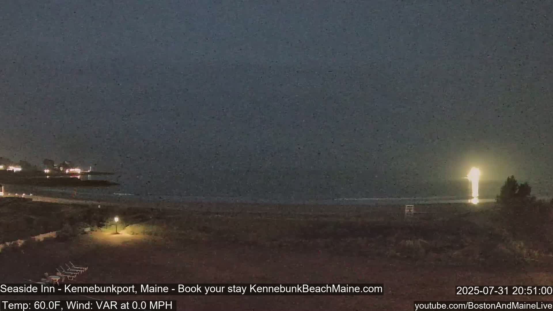 A nighttime view of a dark beach with a brightly lit structure visible in the distance across calm water under a dusky sky.