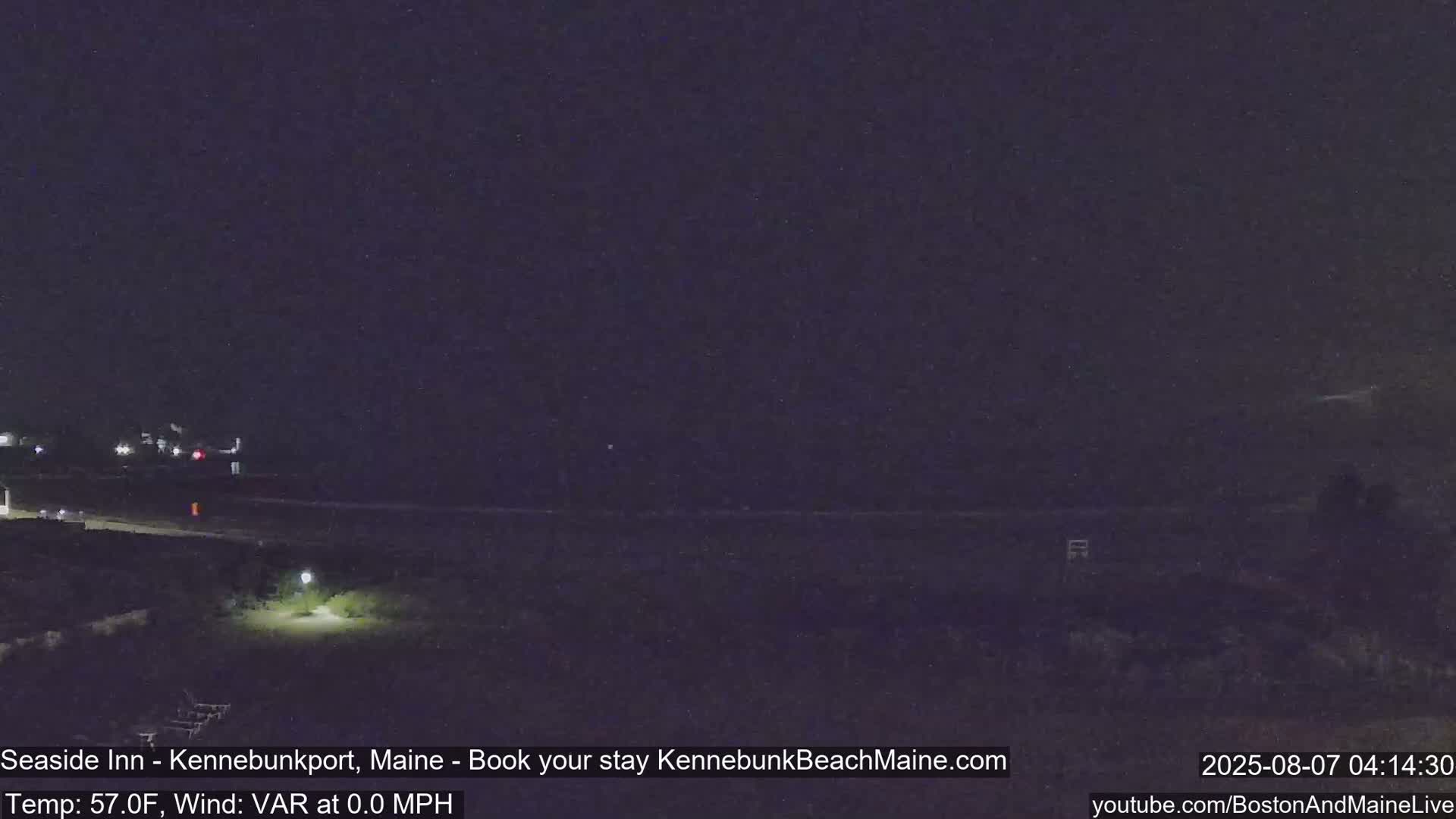 A nighttime view of a dark beach and shoreline with a single lit lamppost in the foreground, under clear skies.