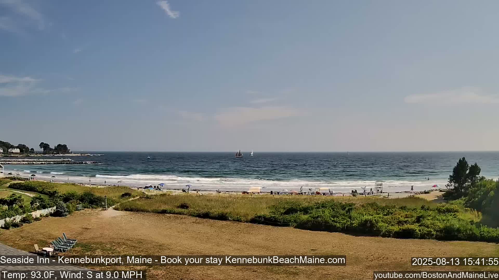 A sandy beach with people and small waves, under a mostly clear sky, is viewed from a grassy dune overlooking the ocean where two sailboats are visible.