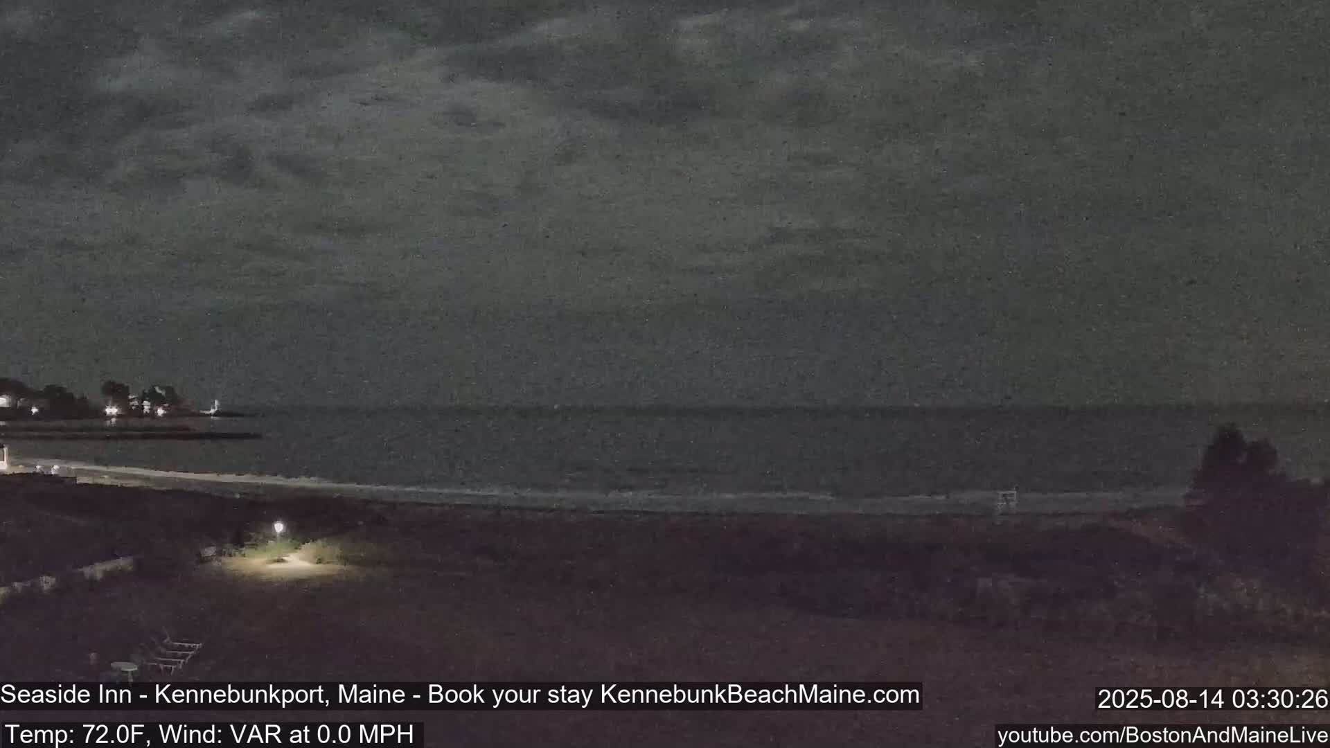 A nighttime view of a dark, calm ocean and sandy beach, under a cloudy sky.