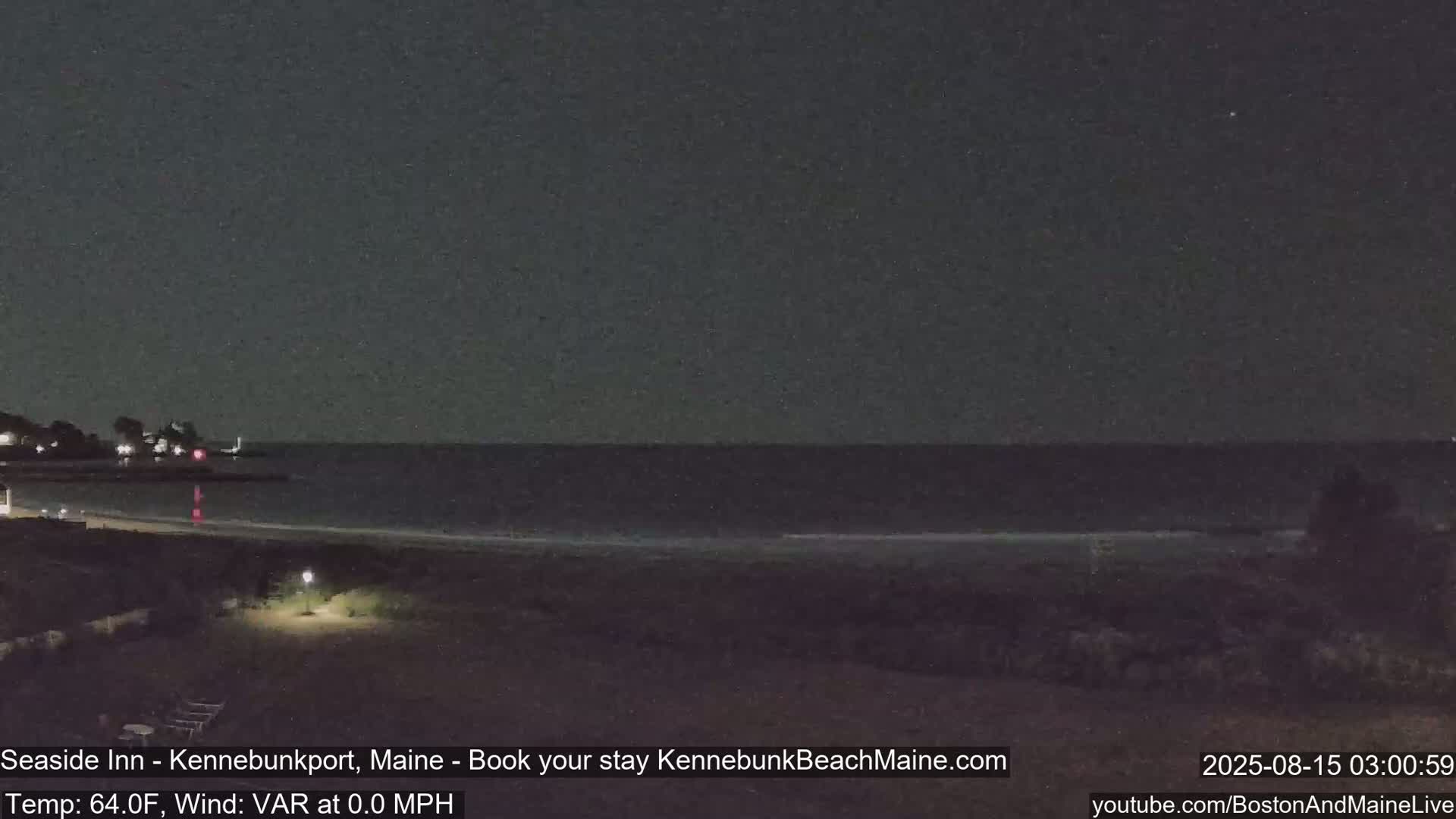 A nighttime view of a calm ocean beach with a dimly lit shoreline and distant lights.