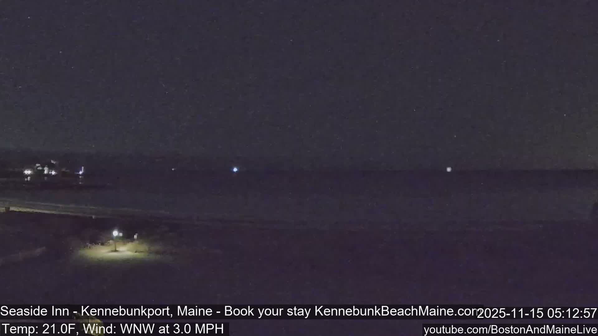 A dark, clear night scene displays a dimly lit beach or path in the foreground, stretching towards a body of water with a few distant lights, under cold conditions of 21.0F and a light WNW wind at 3.0 MPH.