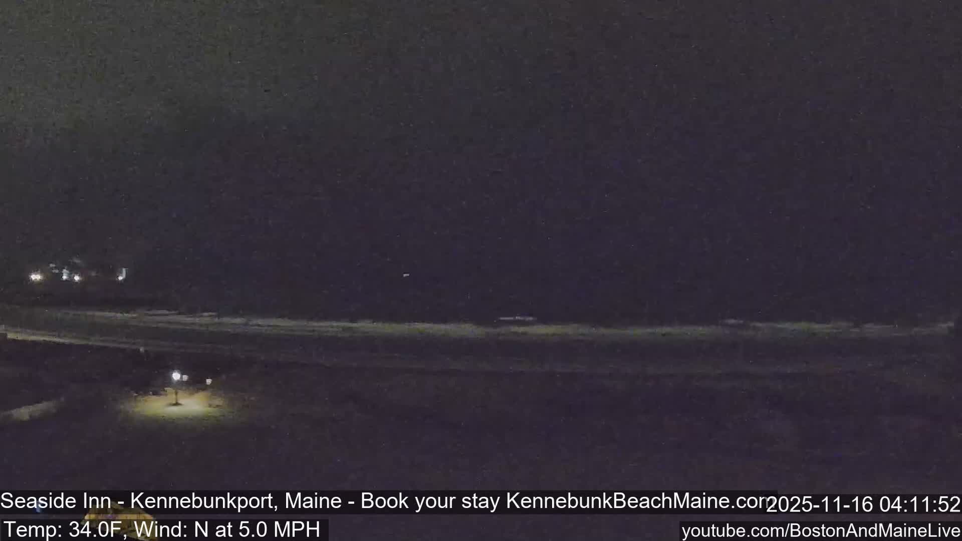 A grainy, dark image captures a beach at night with visible waves breaking on the shore, distant lights from buildings on the left, and a single street lamp illuminating the foreground under a clear sky with a light breeze.
