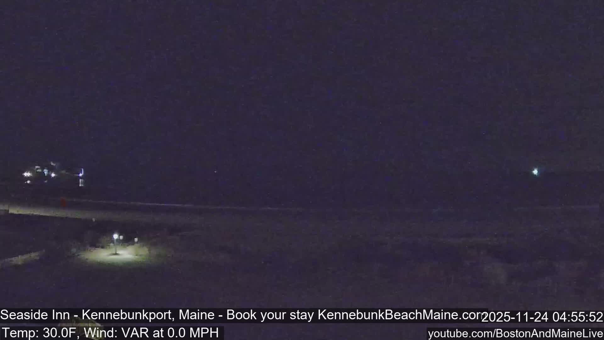 The image shows a clear, dark night at what appears to be a coastal area, with distant building lights on the left and a bright street lamp illuminating a foreground path leading towards a faint horizon.