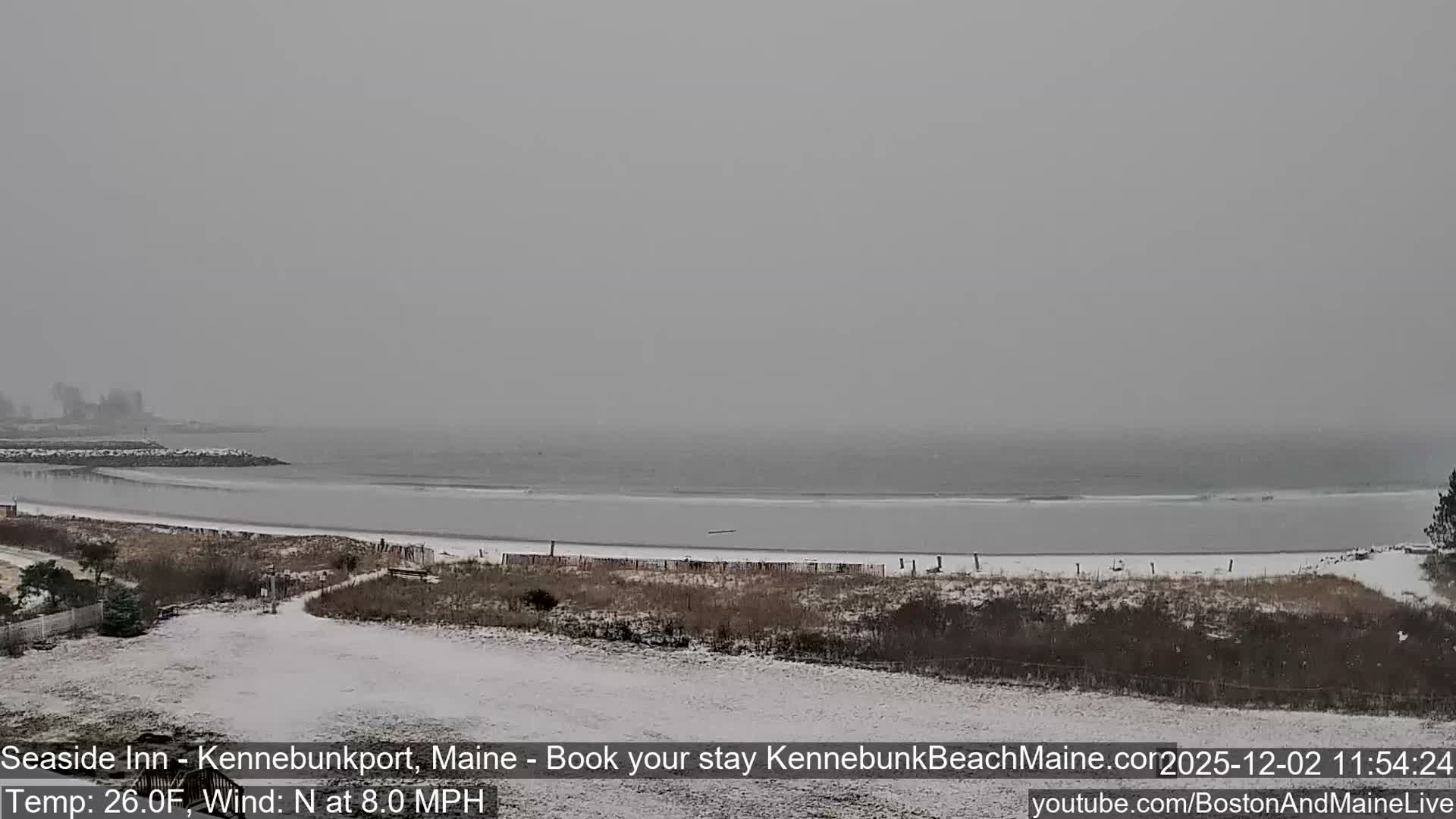 A snow-dusted coastal scene unfolds under a grey, overcast sky, with a choppy ocean stretching to a hazy horizon and a jetty extending from the left shore.