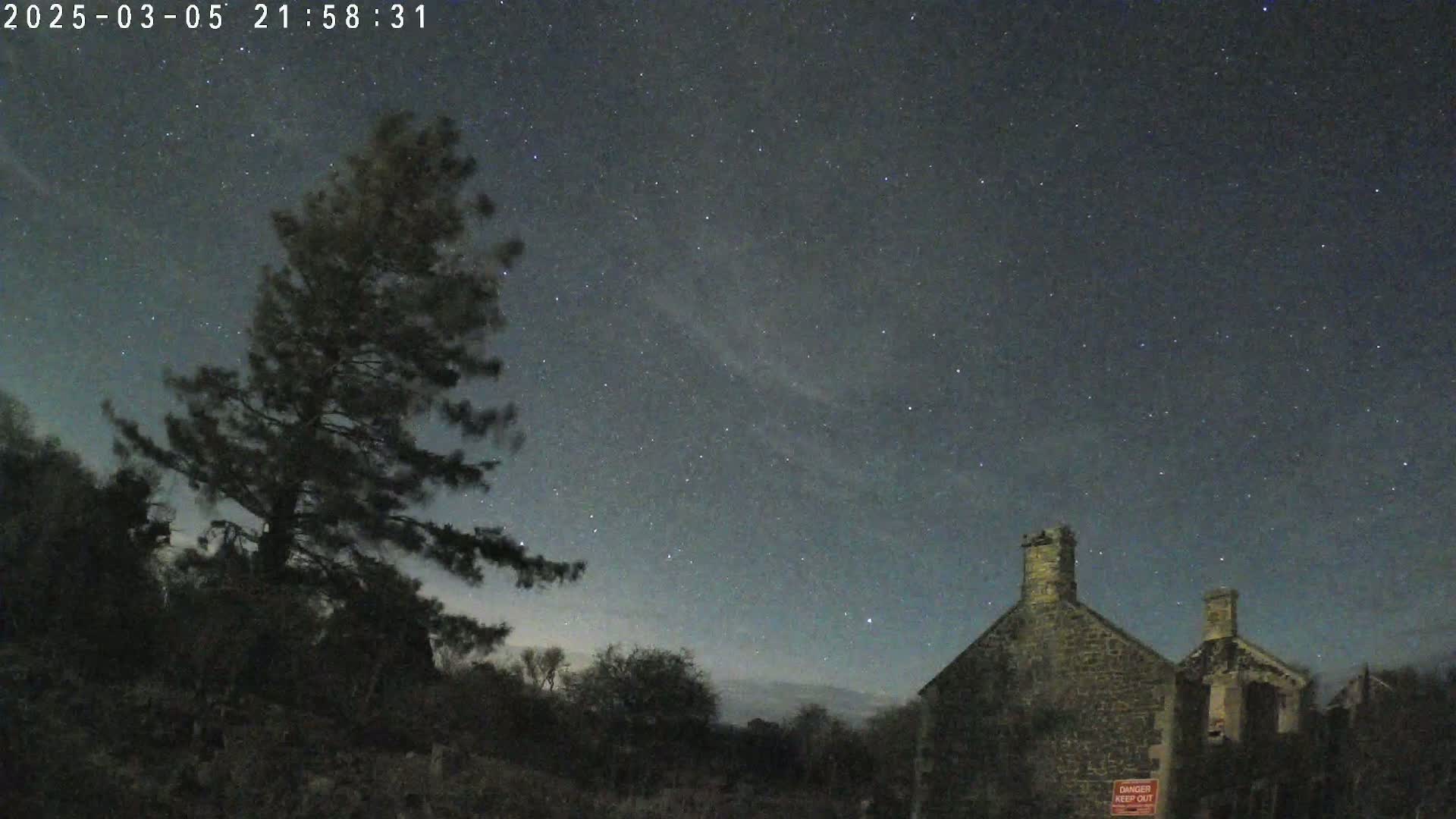 A dark, star-filled night sky overlooks a lone tree and a stone building.