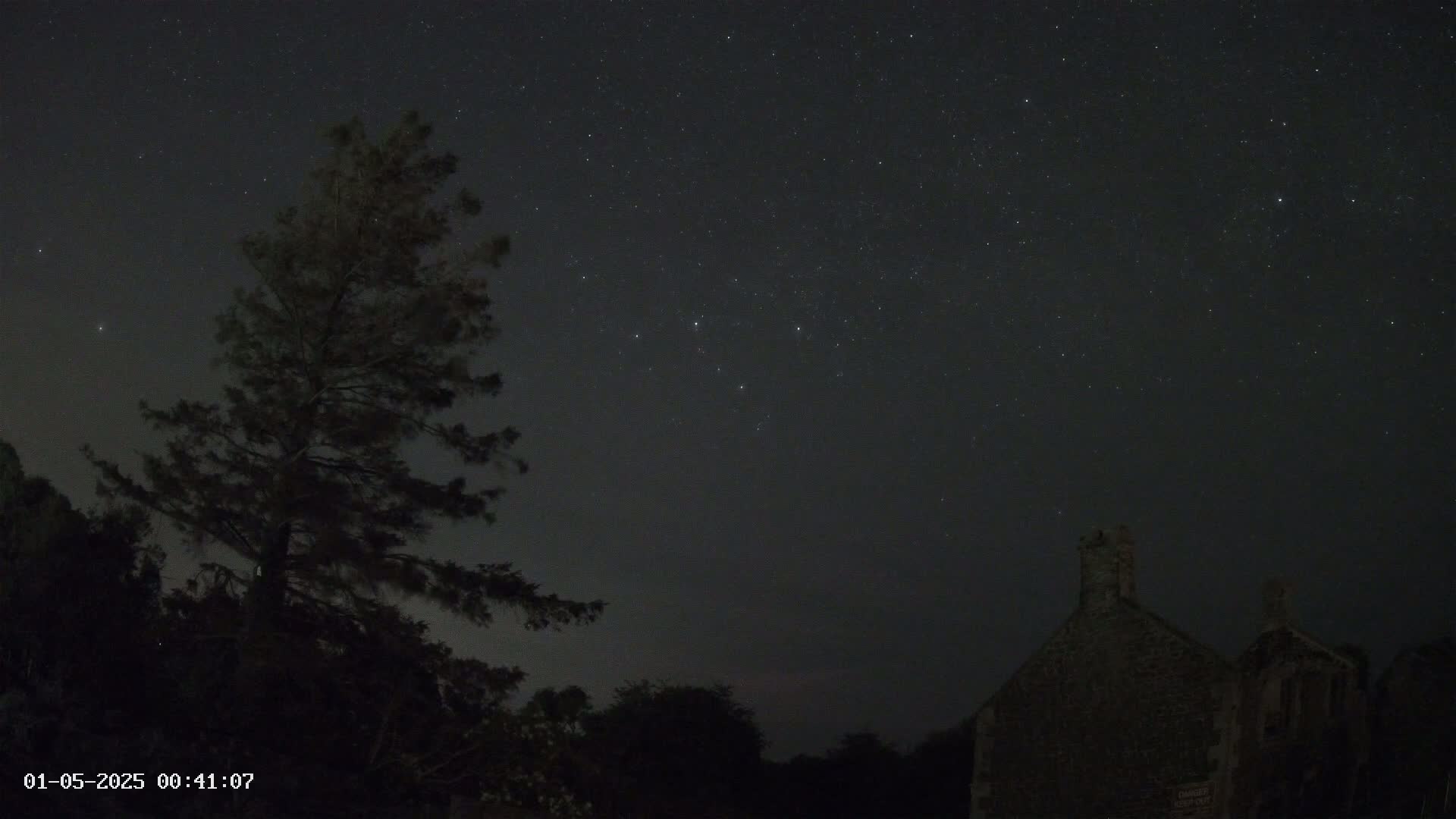 A dark silhouette of a tree and a stone building are visible under a clear, starlit night sky.