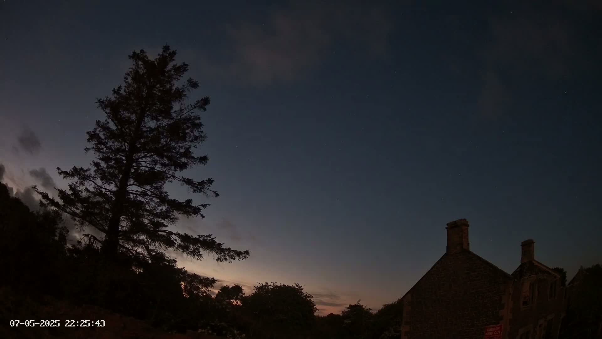 A tall, dark evergreen tree stands to the left of a stone building under a twilight sky dotted with stars and a few clouds.