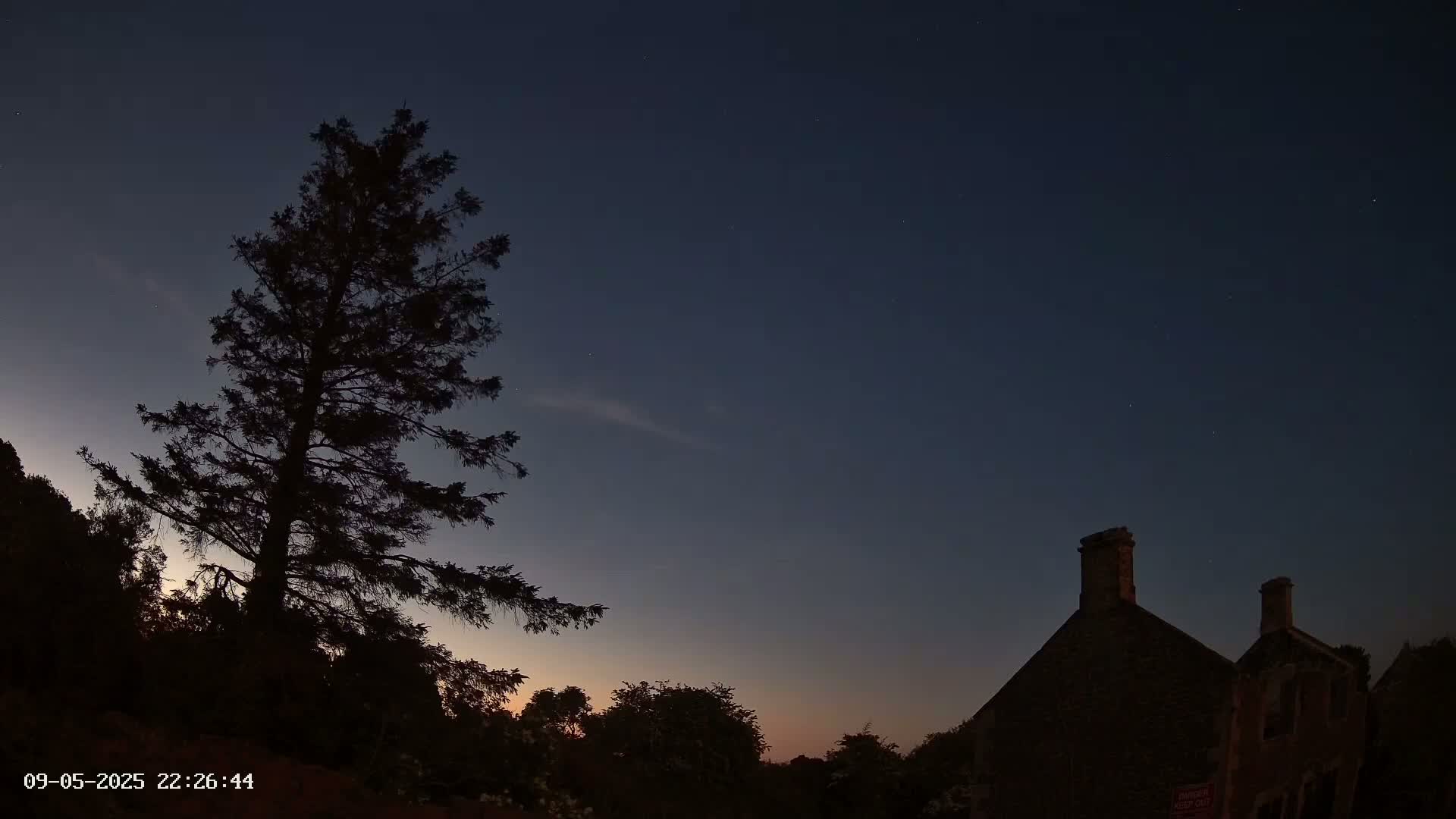Under a dark, twilight sky dotted with stars, a tall, silhouetted evergreen tree stands to the left of a dark, stone building with two chimneys.