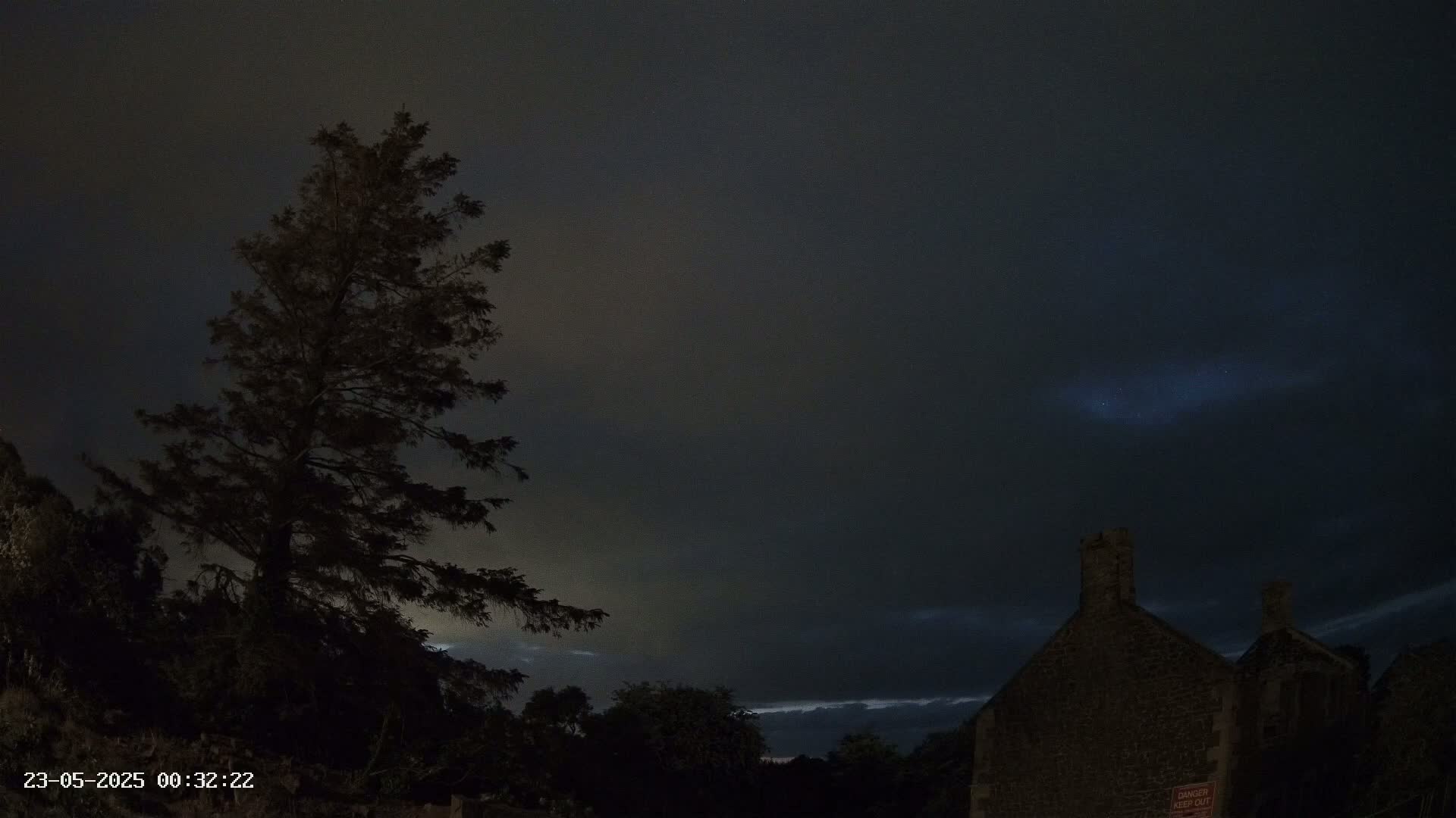 A tall, dark tree stands silhouetted against a night sky partially clouded over, near a stone building under a dark, mostly cloudy sky.
