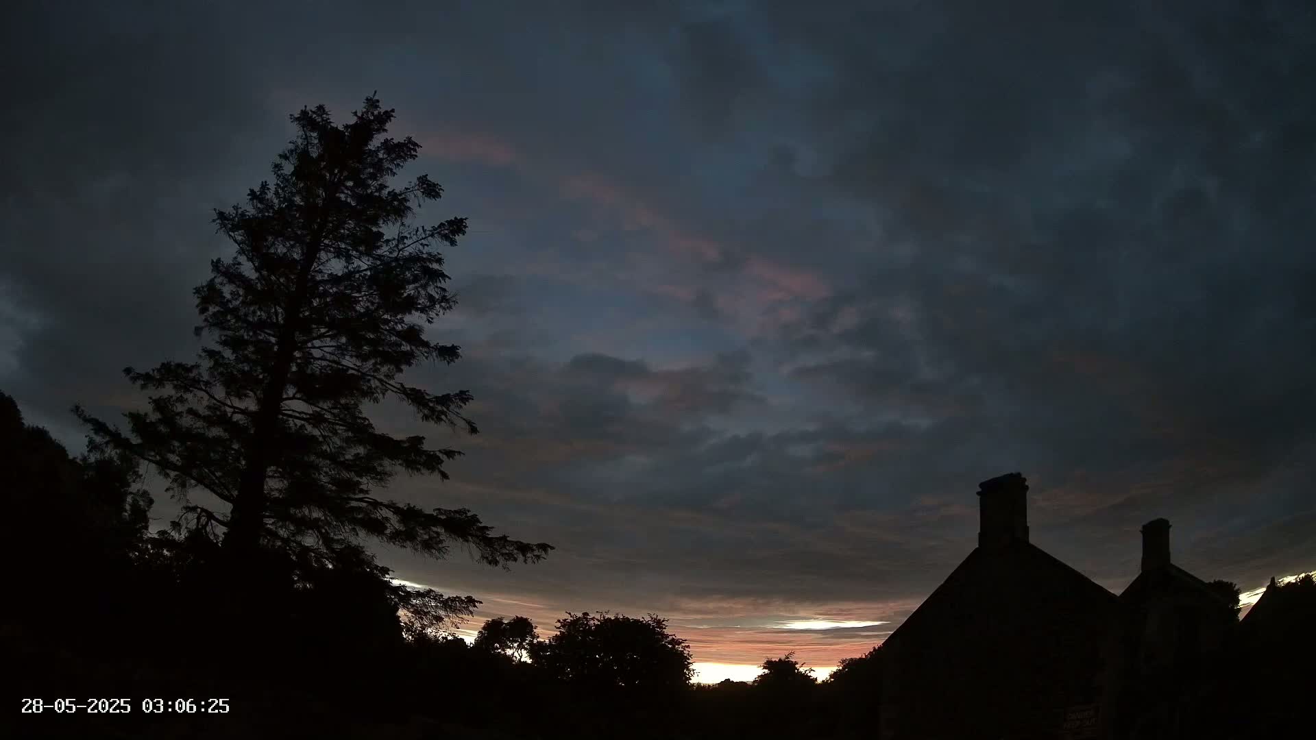 Silhouetted trees and buildings are seen against a dark, cloudy sky with faint streaks of pink and orange near the horizon.
