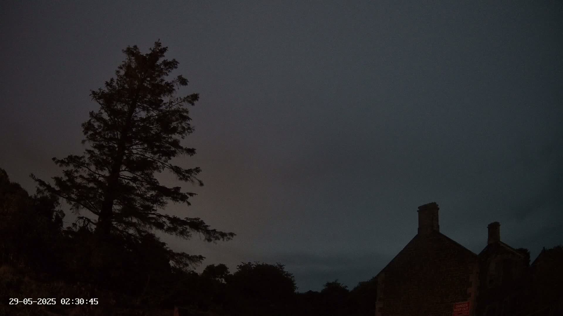 Under a dark, overcast sky, a tall, silhouetted evergreen tree stands to the left of a dark stone building with two chimneys.