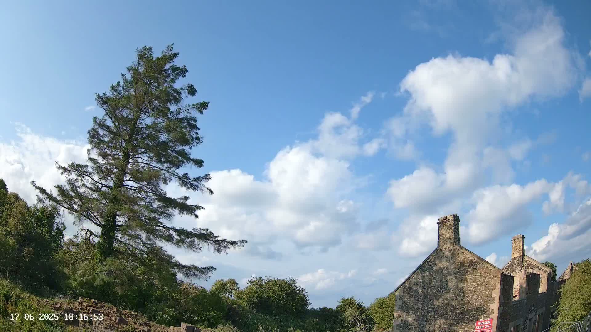 A tall pine tree stands to the left of a partly ruined stone building under a partly cloudy blue sky.