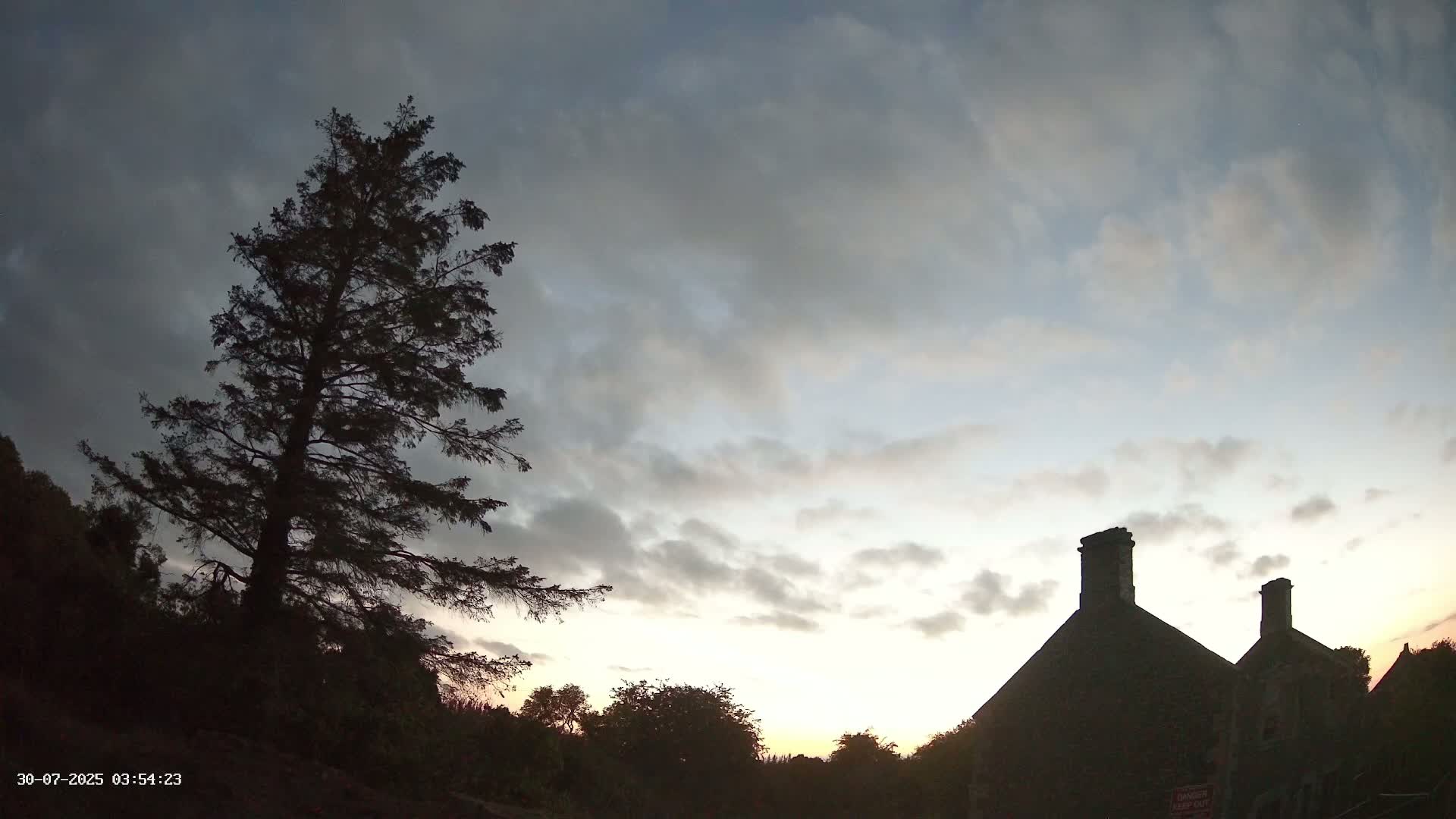 A tall evergreen tree stands to the left of a partially ruined stone building under a mostly clear blue sky with a few wispy clouds.