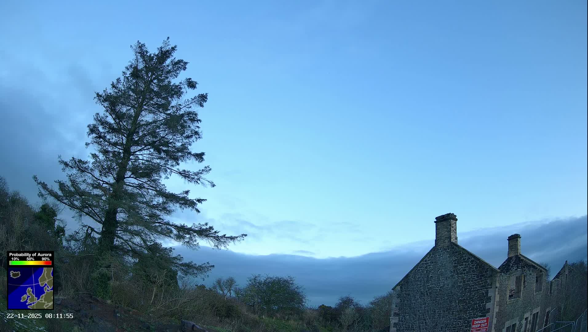 A tall evergreen tree stands on a verdant hillside next to an old stone building, all beneath a mostly overcast sky with hints of clearer blue on the left.