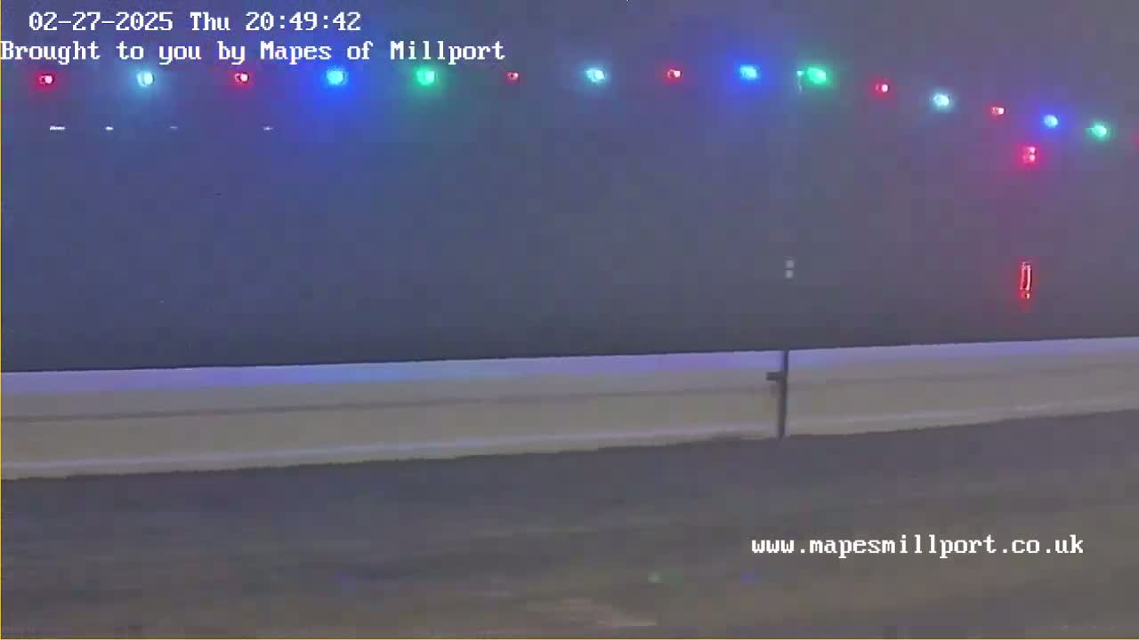 A hazy night scene shows a long, low white barrier in the foreground, with a line of multicolored lights in the distance.