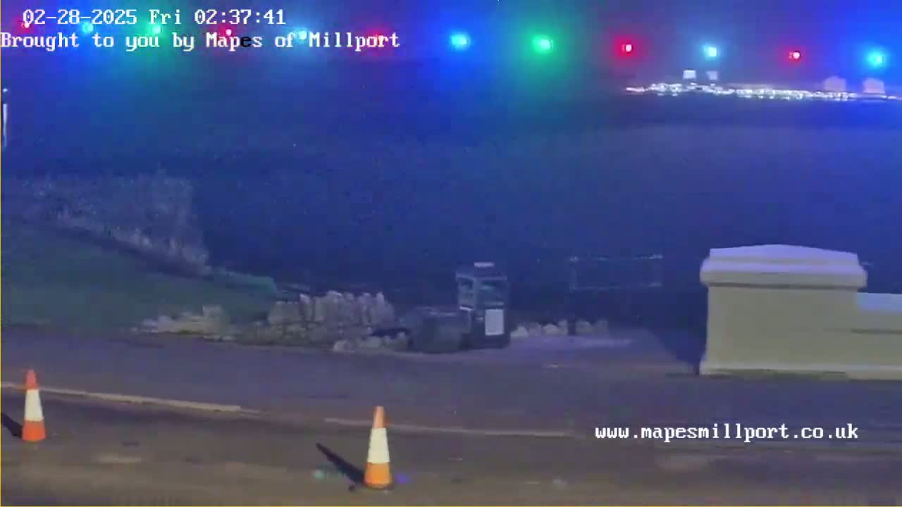 At night, under a dark, hazy sky, a coastal scene shows a road with traffic cones, a low stone wall, and a distant, faintly lit town across a body of water.