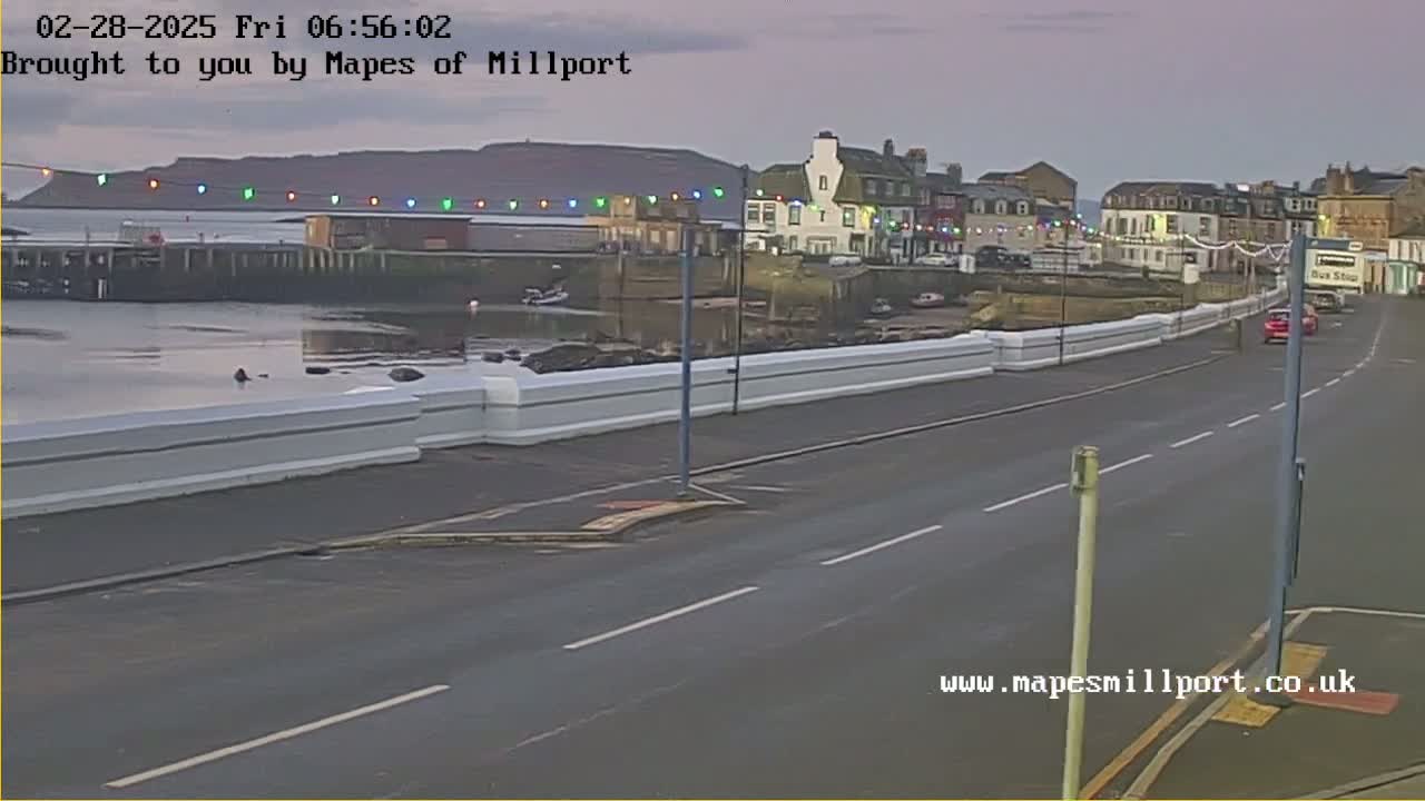A seaside town at dawn, with a calm body of water and a road running alongside a low white wall, under a dusky sky.