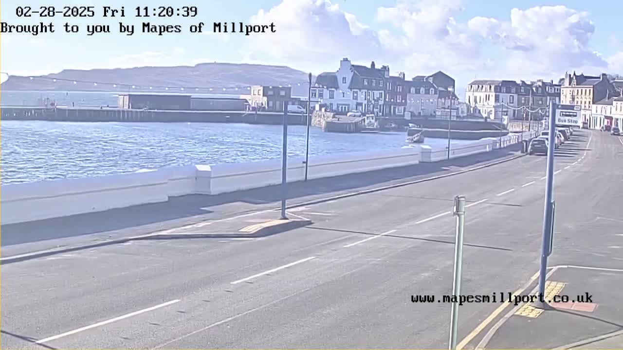 A mostly sunny day shows a seaside town with a road alongside a waterfront, featuring parked cars and buildings.