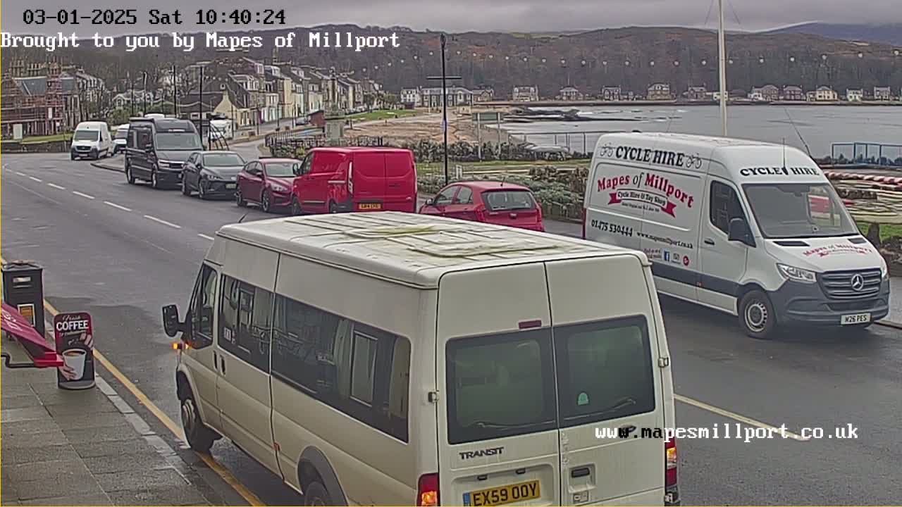 Several vehicles are parked and driving on a street near a coastline under an overcast sky.