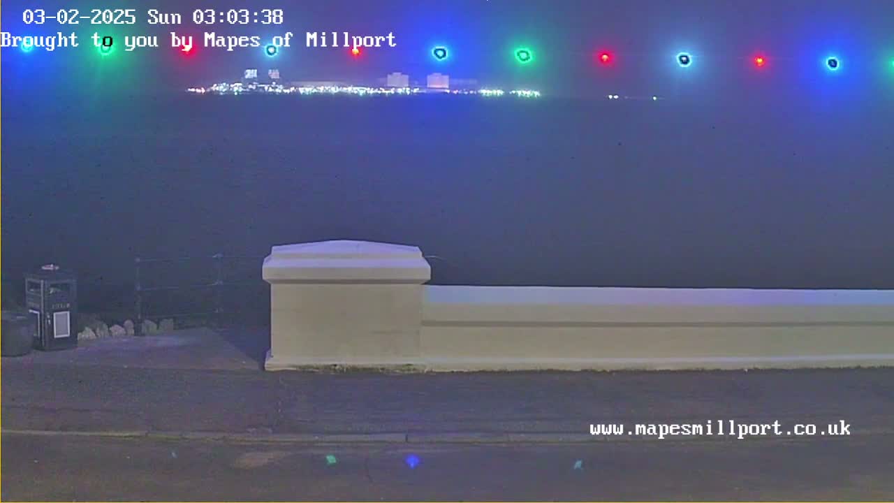 A nighttime view of a body of water with lights on the opposite shore, seen from behind a low white wall under hazy conditions.