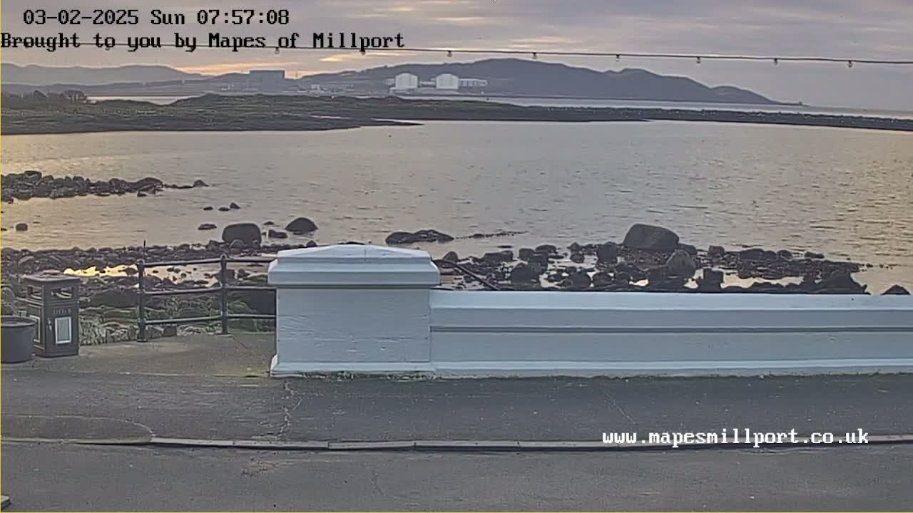 A calm body of water with dark rocks lining the shore is viewed from a paved area with a low white wall at sunrise.