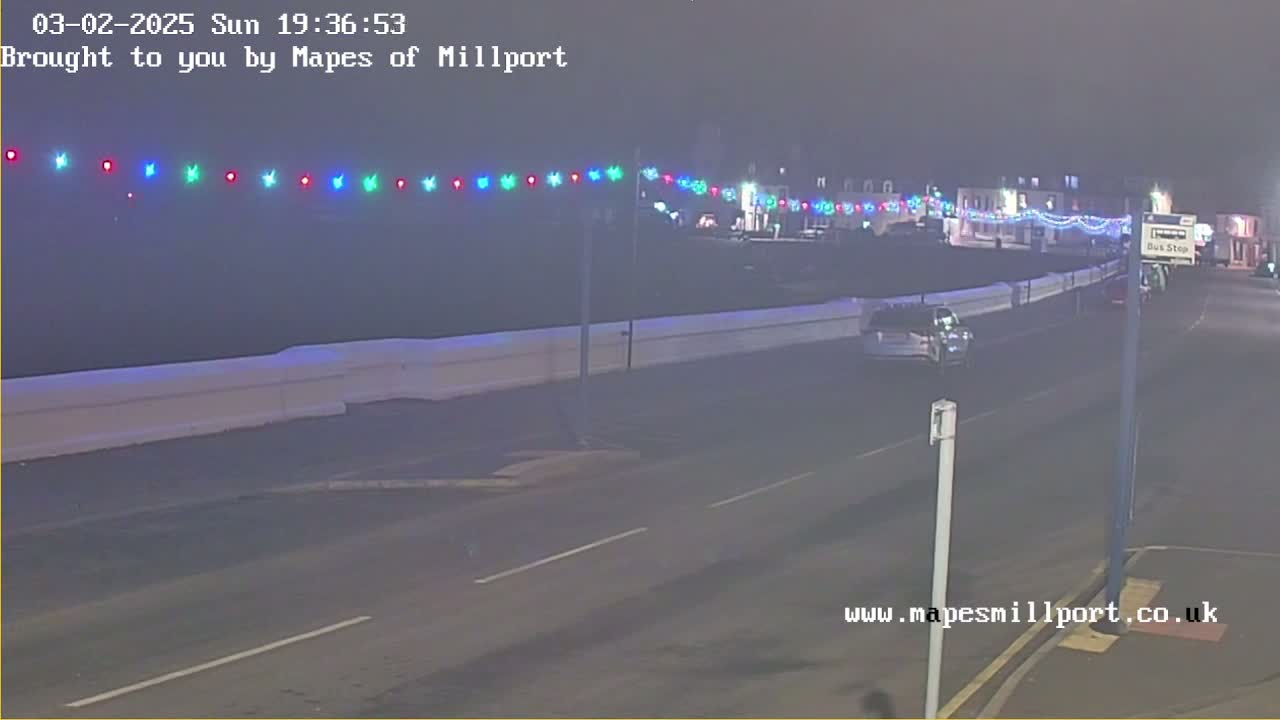 A nighttime street scene shows a road with a low white barrier separating it from a beachfront area, decorated with colorful string lights, and a few cars are visible.