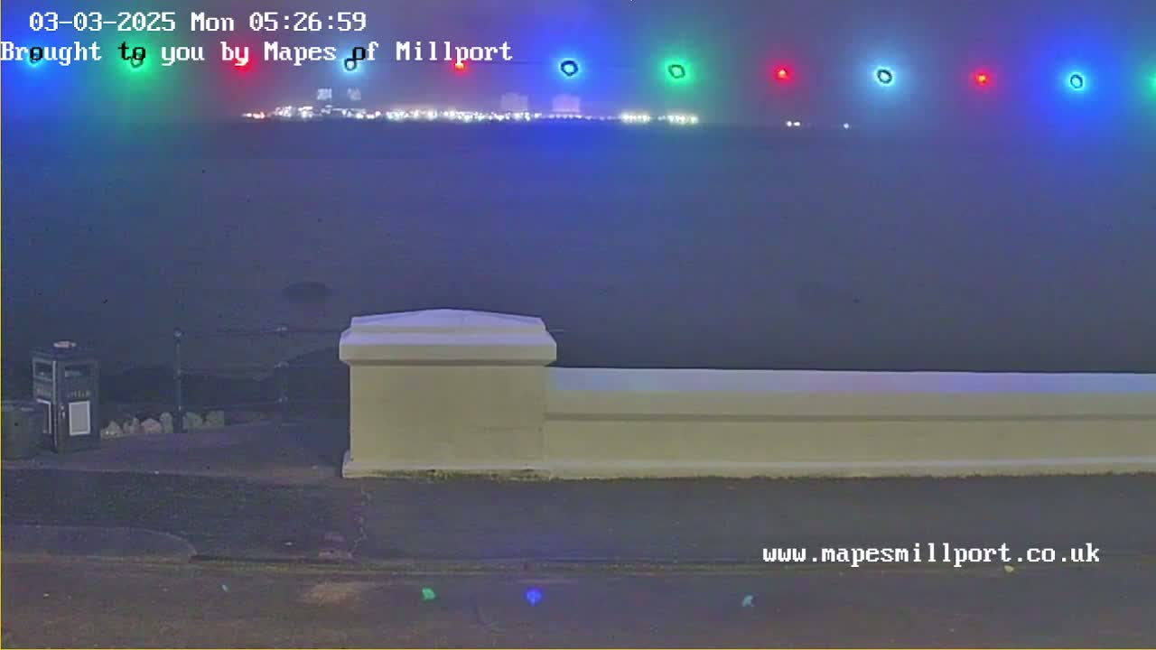 A nighttime view across a body of water toward a distant, brightly lit town, partially obscured by fog, seen from behind a low white wall.