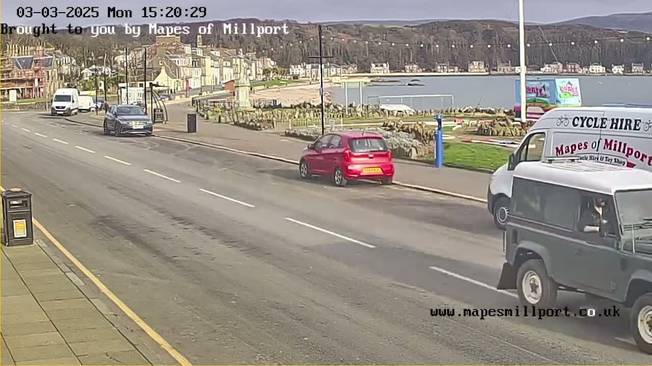 Several cars are parked and driving on a road next to a seaside town on a sunny day.