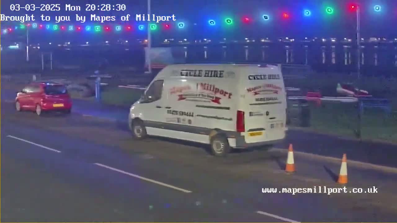 A red car and a white van are driving on a road at night under hazy conditions, with traffic lights visible in the background.