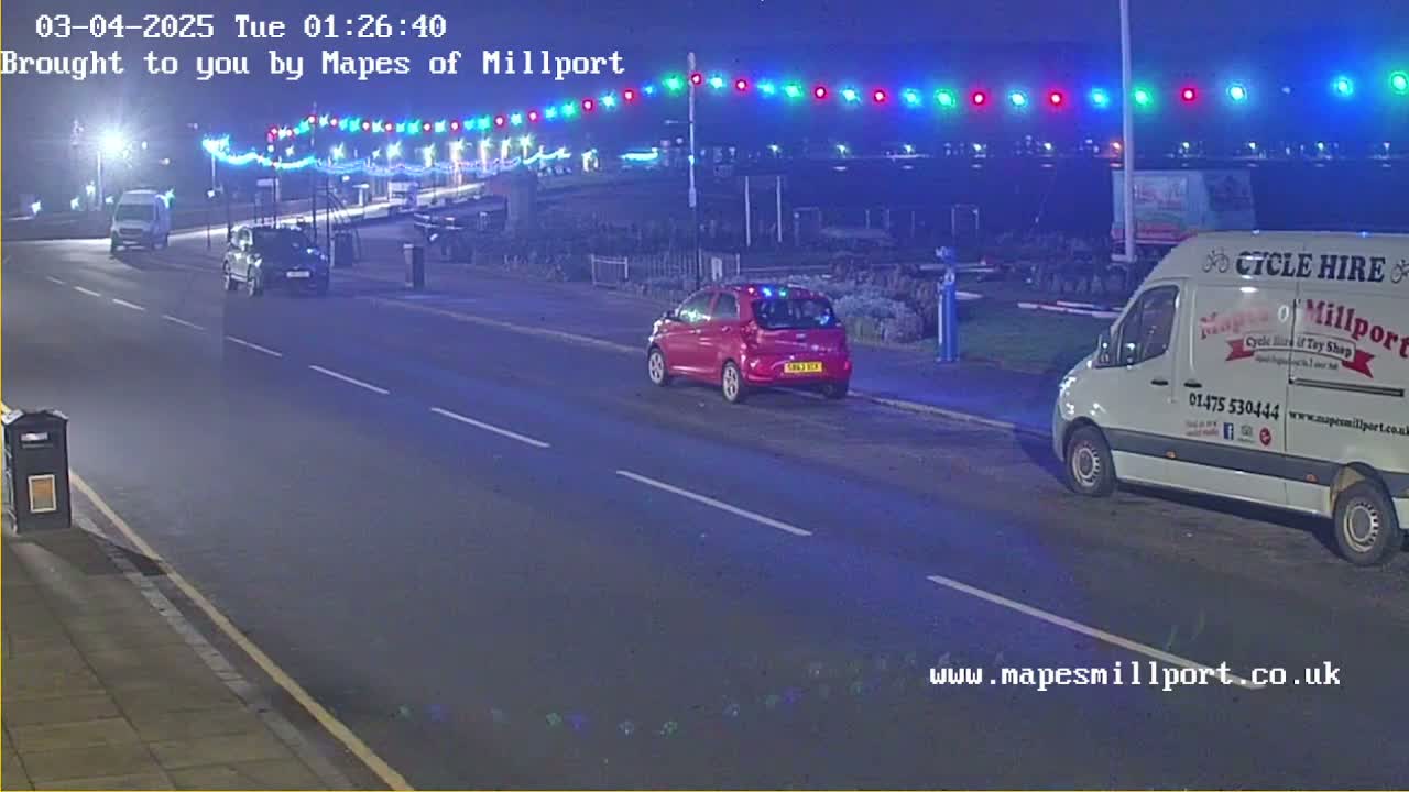 At night, a few cars are parked or driving on a mostly empty road lined with colorful lights, under a clear sky.