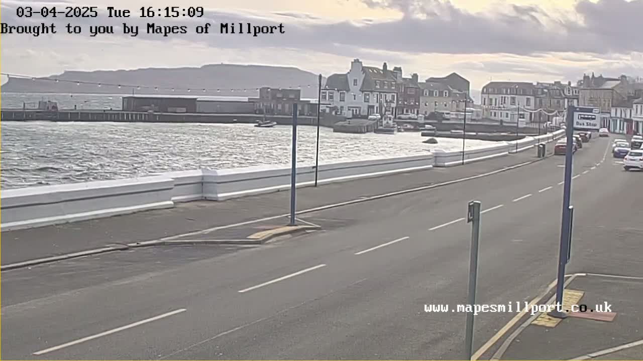 A windy, overcast day shows a waterfront town with a road and parked cars, a low white wall separating the road from the choppy water.