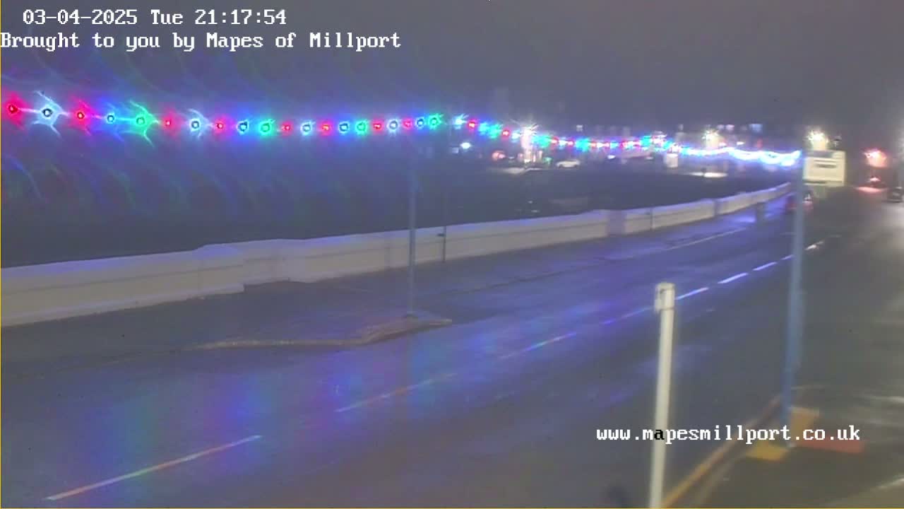 A wet, dark road at night is illuminated by multicolored lights strung across a low wall.