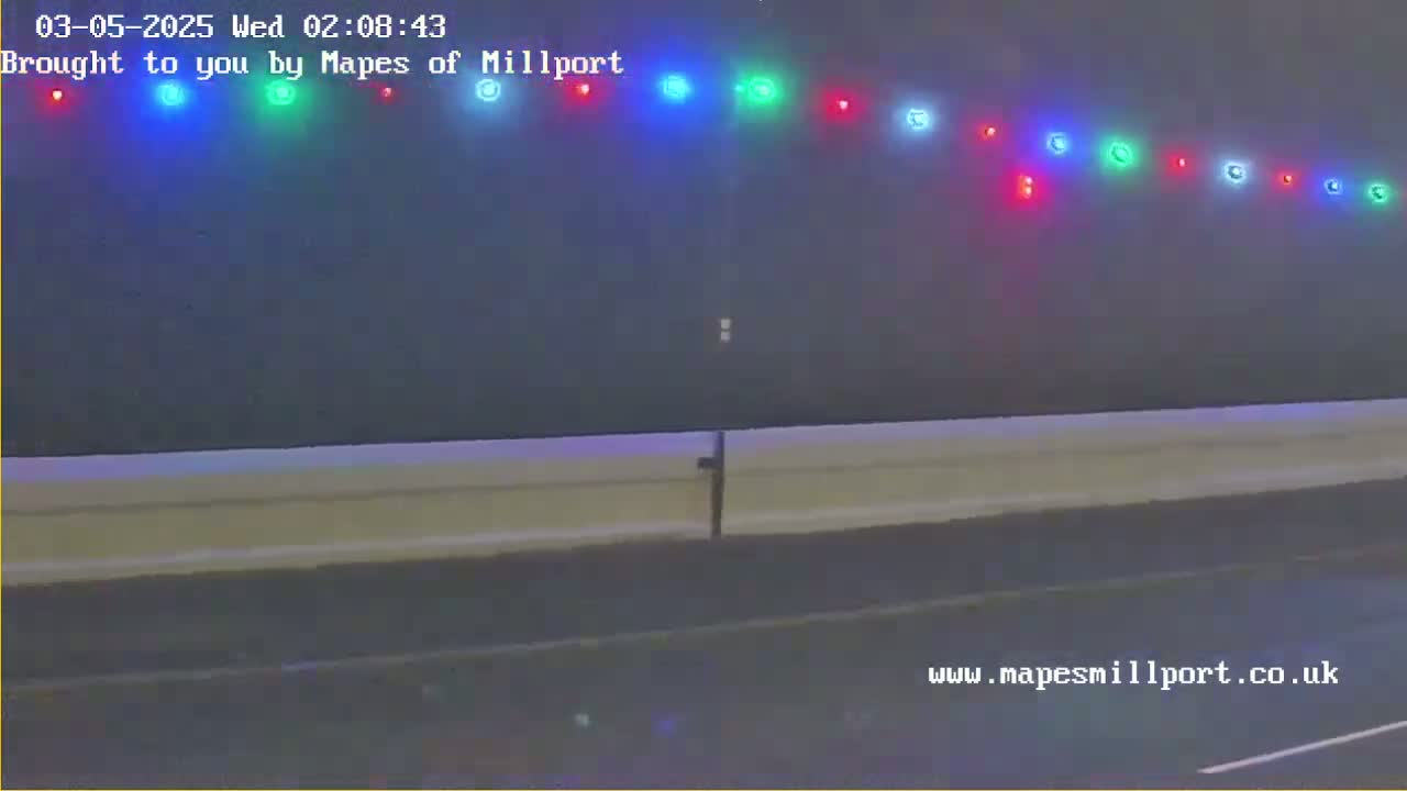 A nighttime, possibly wet, view of a road with a low wall and a line of multicolored lights above it.