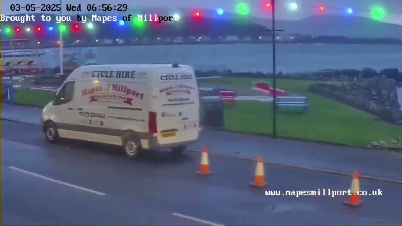 A white van is parked on a wet road next to two orange traffic cones, with a body of water and distant hills in the background under a dark, overcast sky.