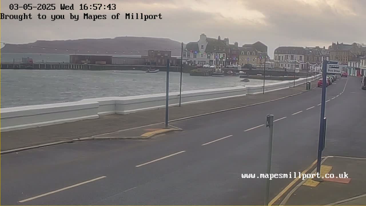 A seaside town is seen on an overcast day, with a choppy body of water in the background and a road with cars parked along the side.
