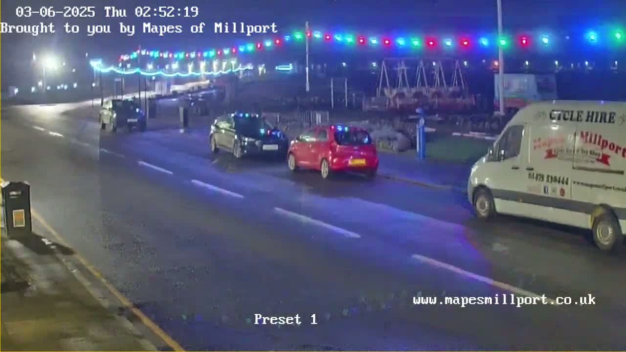 Three vehicles are traveling on a wet road at night under a string of multicolored lights.