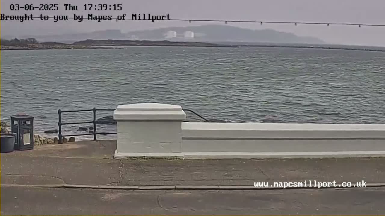 A slightly overcast day shows a calm body of water behind a low white wall and paved walkway; a trash receptacle is visible near the wall.