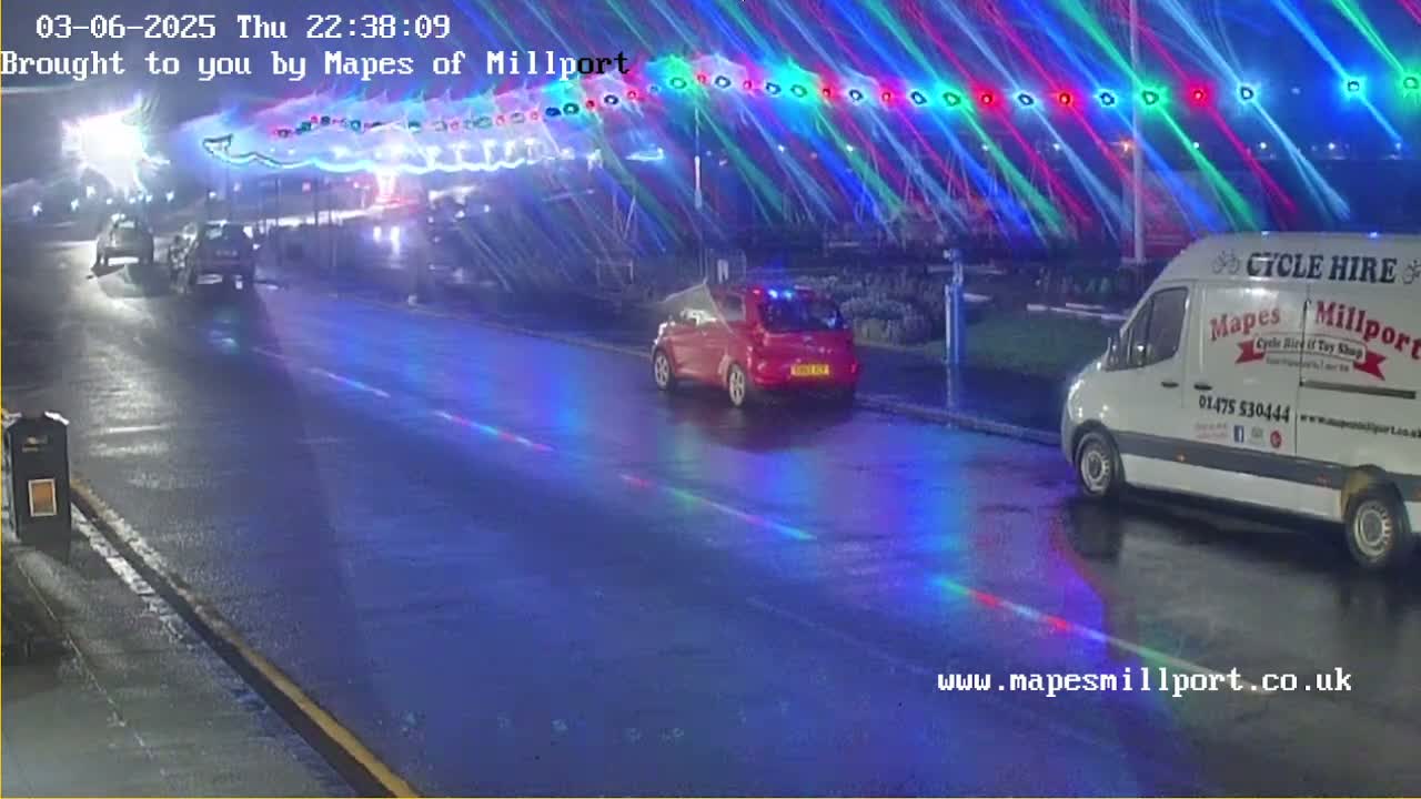 Several cars are parked on a wet road at night under multicolored light beams.