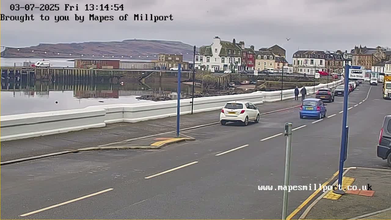 A seaside town on an overcast day, with several cars parked and driving along a road next to a harbor wall.