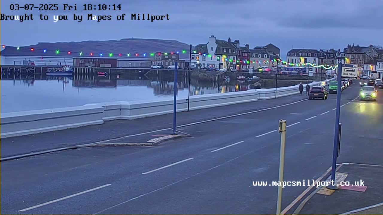 A twilight view shows a calm body of water reflecting a waterfront town with colorful lights strung across buildings, and a road with several cars and pedestrians.